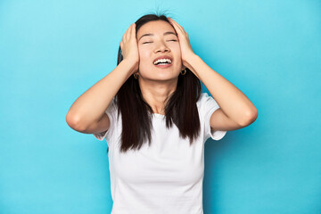 Wall Mural - Young Asian woman in white t-shirt, studio shot, laughs joyfully keeping hands on head. Happiness concept.