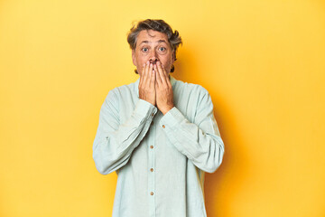 Wall Mural - Middle-aged man posing on a yellow backdrop shocked, covering mouth with hands, anxious to discover something new.