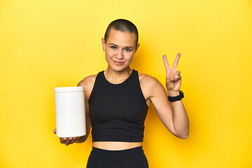 Sportswoman with protein shake, yellow studio background showing number two with fingers.