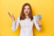 Redhead with dollar bills, cash in hand receiving a pleasant surprise, excited and raising hands.