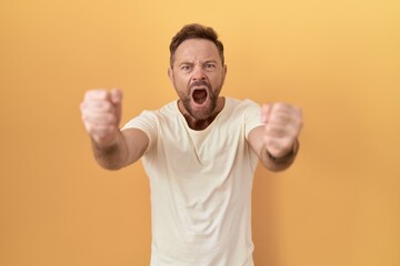 Poster - Middle age man with beard standing over yellow background angry and mad raising fists frustrated and furious while shouting with anger. rage and aggressive concept.