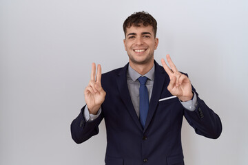 Wall Mural - Young hispanic business man wearing suit and tie smiling looking to the camera showing fingers doing victory sign. number two.