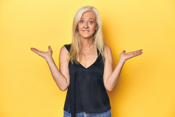 Elegant middle-aged woman in black top, yellow studio confused and doubtful shrugging shoulders to hold a copy space.