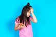 Upset depressed Caucasian kid girl wearing pink t-shirt over blue background makes face palm as forgot about something important holds mobile phone expresses sorrow and regret blames