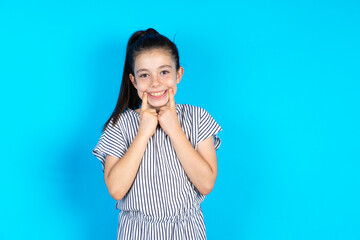 Happy Caucasian kid girl wearing striped dress over blue background with toothy smile, keeps index fingers near mouth, fingers pointing and forcing cheerful smile