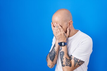 Poster - Hispanic man with tattoos standing over blue background with sad expression covering face with hands while crying. depression concept.