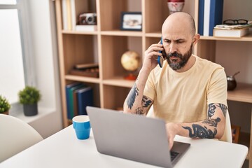 Sticker - Young bald man using laptop talking on smartphone at home