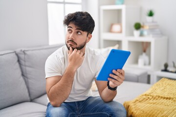 Wall Mural - Hispanic man with beard using touchpad sitting on the sofa with hand on chin thinking about question, pensive expression. smiling with thoughtful face. doubt concept.