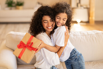 Mother's day concept. Adorable latin girl giving gift and embracing mom, mommy and daughter bonding together at home, sitting on sofa