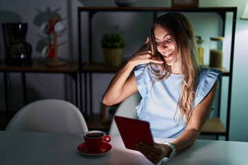 Sticker - Young hispanic woman using touchpad sitting on the table at night smiling doing phone gesture with hand and fingers like talking on the telephone. communicating concepts.