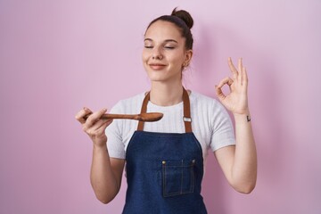 Canvas Print - Young hispanic girl wearing professional cook apron holding wood spoon winking looking at the camera with sexy expression, cheerful and happy face.