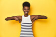 © Asier - Vibrant African woman, white tank-top, yellow studio, points down with fingers, positive feeling.