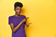 © Asier - African woman in violet tee, yellow studio scene, shocked pointing with index fingers to a copy space.