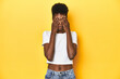 © Asier - African woman, white tee, short hair, yellow studio, afraid covering eyes with hands.
