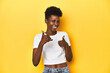 © Asier - African woman, white tee, short hair, yellow studio, raising both thumbs up, smiling and confident.