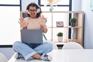 Poster - Young hispanic woman using laptop sitting on the table wearing headphones afraid and terrified with fear expression stop gesture with hands, shouting in shock. panic concept.