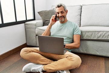 Wall Mural - Middle age grey-haired man talking on smartphone using laptop sitting on floor at home