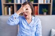 © Krakenimages.com - Middle age hispanic woman sitting on the sofa at home peeking in shock covering face and eyes with hand, looking through fingers with embarrassed expression.