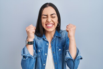 Wall Mural - Hispanic woman standing over blue background excited for success with arms raised and eyes closed celebrating victory smiling. winner concept.