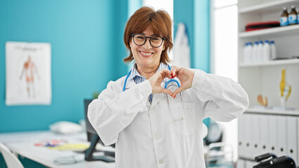 Wall Mural - Middle age woman doctor smiling doing heart gesture with hands at clinic