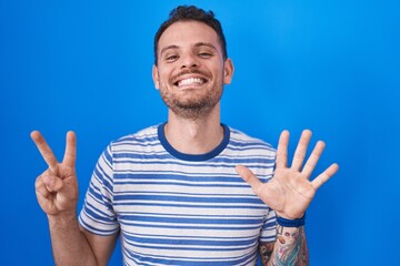 Sticker - Young hispanic man standing over blue background showing and pointing up with fingers number seven while smiling confident and happy.