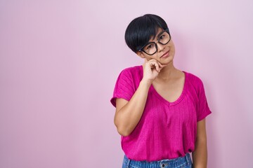 Poster - Young asian woman with short hair standing over pink background with hand on chin thinking about question, pensive expression. smiling with thoughtful face. doubt concept.