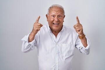 Canvas Print - Senior man with grey hair standing over isolated background smiling amazed and surprised and pointing up with fingers and raised arms.