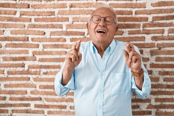 Sticker - Senior man with grey hair standing over bricks wall gesturing finger crossed smiling with hope and eyes closed. luck and superstitious concept.