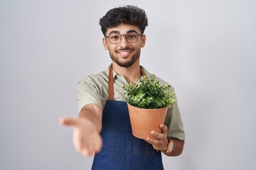 Arab man with beard holding green plant pot smiling cheerful offering palm hand giving assistance and acceptance.