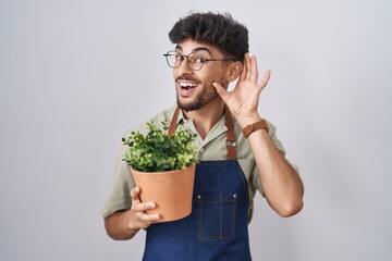 Poster - Arab man with beard holding green plant pot smiling with hand over ear listening an hearing to rumor or gossip. deafness concept.