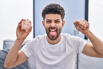 Poster - Arab man holding piggy bank and house keys sticking tongue out happy with funny expression.