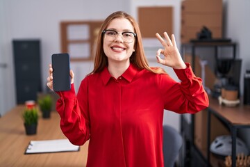 Canvas Print - Redhead woman working at the office showing smartphone screen doing ok sign with fingers, smiling friendly gesturing excellent symbol