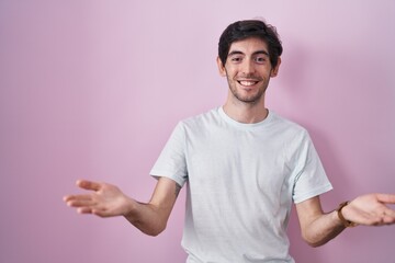 Wall Mural - Young hispanic man standing over pink background smiling cheerful offering hands giving assistance and acceptance.