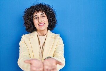 Poster - Young brunette woman with curly hair standing over blue background smiling with hands palms together receiving or giving gesture. hold and protection