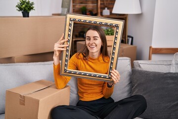 Poster - Young caucasian woman at new home holding empty frame winking looking at the camera with sexy expression, cheerful and happy face.