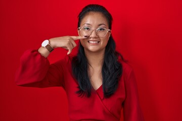 Poster - Asian young woman standing over red background pointing with hand finger to face and nose, smiling cheerful. beauty concept