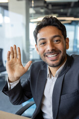 Wall Mural - Vertical shot, young successful businessman looking into smartphone camera talking on video call using app on phone, smiling and having friendly conversation with friends and colleague inside office.