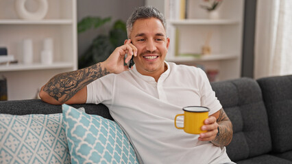 Sticker - Young hispanic man talking on smartphone drinking coffee at home