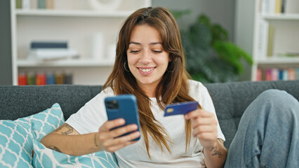 Wall Mural - Young beautiful hispanic woman shopping with smartphone and credit card sitting on sofa at home