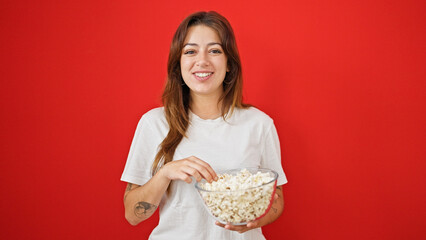 Wall Mural - Young beautiful hispanic woman eating popcorn smiling over isolated red background