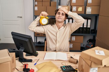 Young man working at small business ecommerce holding alarm clock stressed and frustrated with hand on head, surprised and angry face