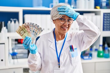 Poster - Middle age woman with grey hair working at scientist laboratory holding polish zloty banknotes stressed and frustrated with hand on head, surprised and angry face