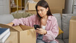 © Krakenimages.com - Young beautiful hispanic woman using smartphone unpacking cardboard box at new home