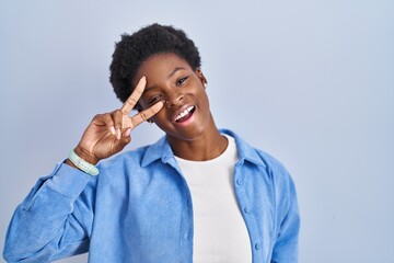 Wall Mural - African american woman standing over blue background doing peace symbol with fingers over face, smiling cheerful showing victory