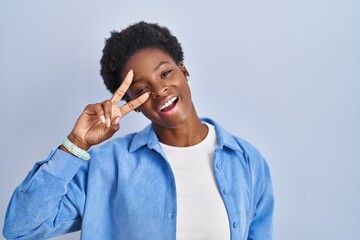 Canvas Print - African american woman standing over blue background doing peace symbol with fingers over face, smiling cheerful showing victory