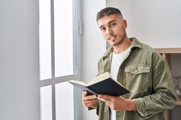 Wall Mural - Young hispanic man reading book standing at home