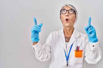 Canvas Print - Middle age woman with grey hair wearing scientist robe amazed and surprised looking up and pointing with fingers and raised arms.