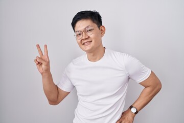 Sticker - Young asian man standing over white background smiling looking to the camera showing fingers doing victory sign. number two.