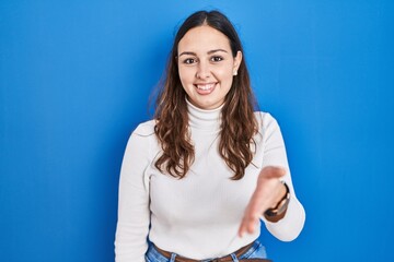 Sticker - Young hispanic woman standing over blue background smiling friendly offering handshake as greeting and welcoming. successful business.