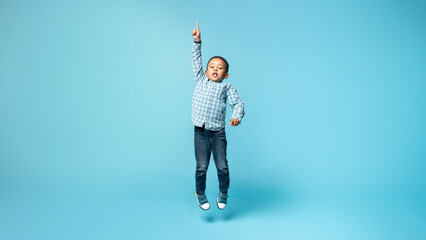 Wall Mural - Little african american child boy jumping and pointing up with finger, blue studio background, panorama with free space